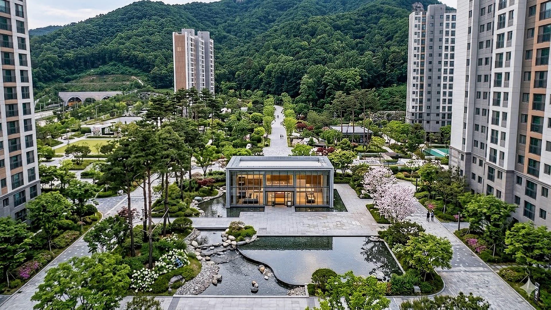 단지 중앙의 물이 흐르는 수경 시설 옆에 세련된 디자인의 티하우스가 위치하고 그 뒤로 필봉산의 쾌적한 숲길이 이어지는 북오산자이 드포레 단지 조경