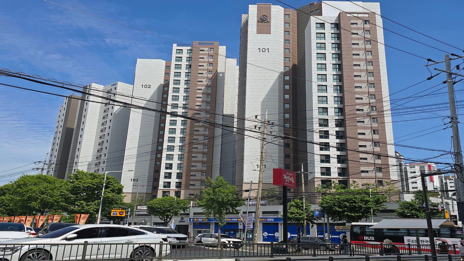 오목천역 더리브 101동, 102동 건물이 푸른 하늘 아래 웅장하게 서 있고 전면에 복잡한 전신주 배선이 지나가는 도로변 외관 전경