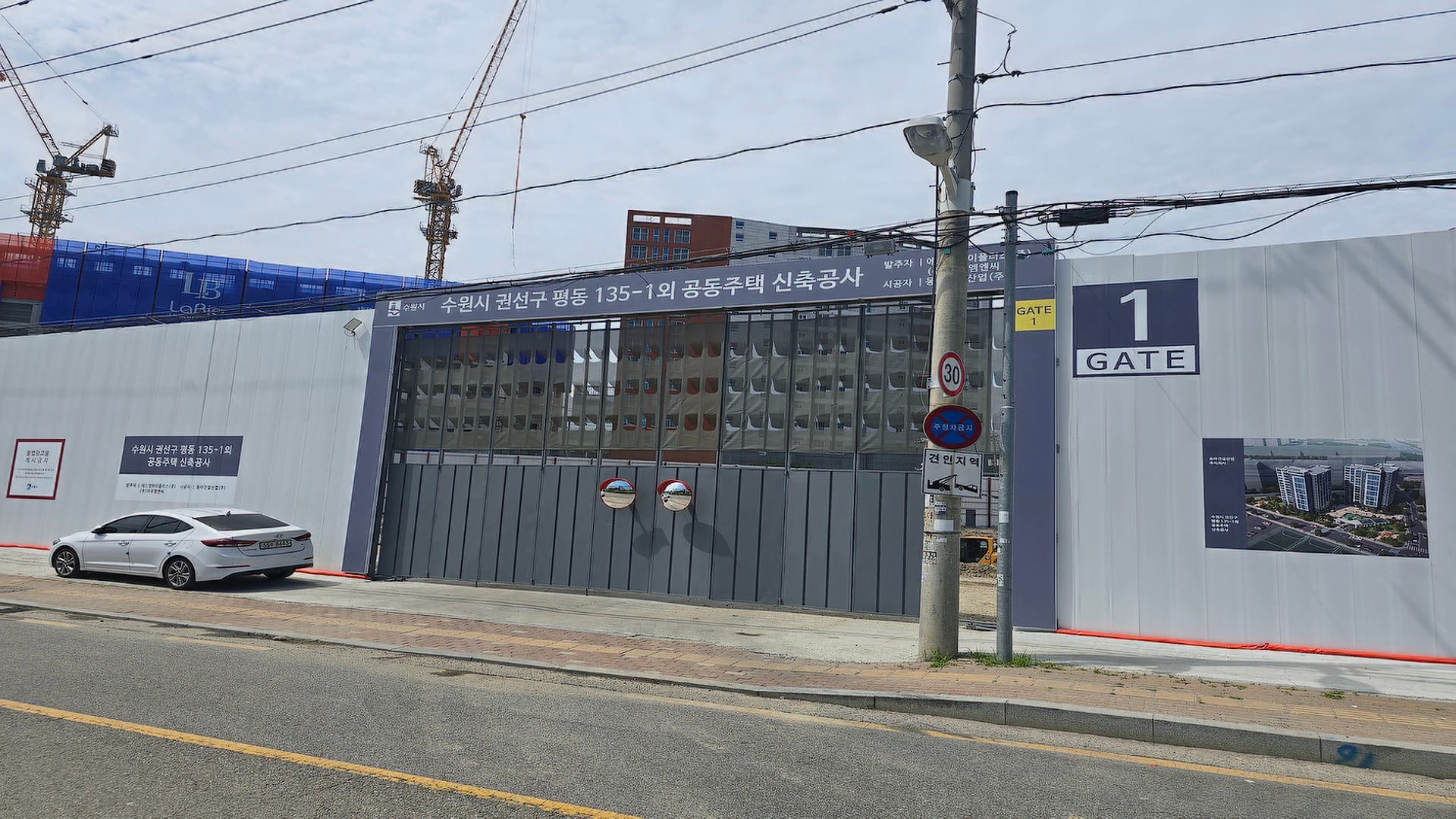수원역 아너스빌 타임원 아파트 공사 현장의 하얀색 가설 펜스와 1번 차량 출입문 및 상단 타워크레인 전경